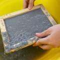 Zwei Hände schöpfen Papierbrei in Rahmen in gelber Wanne